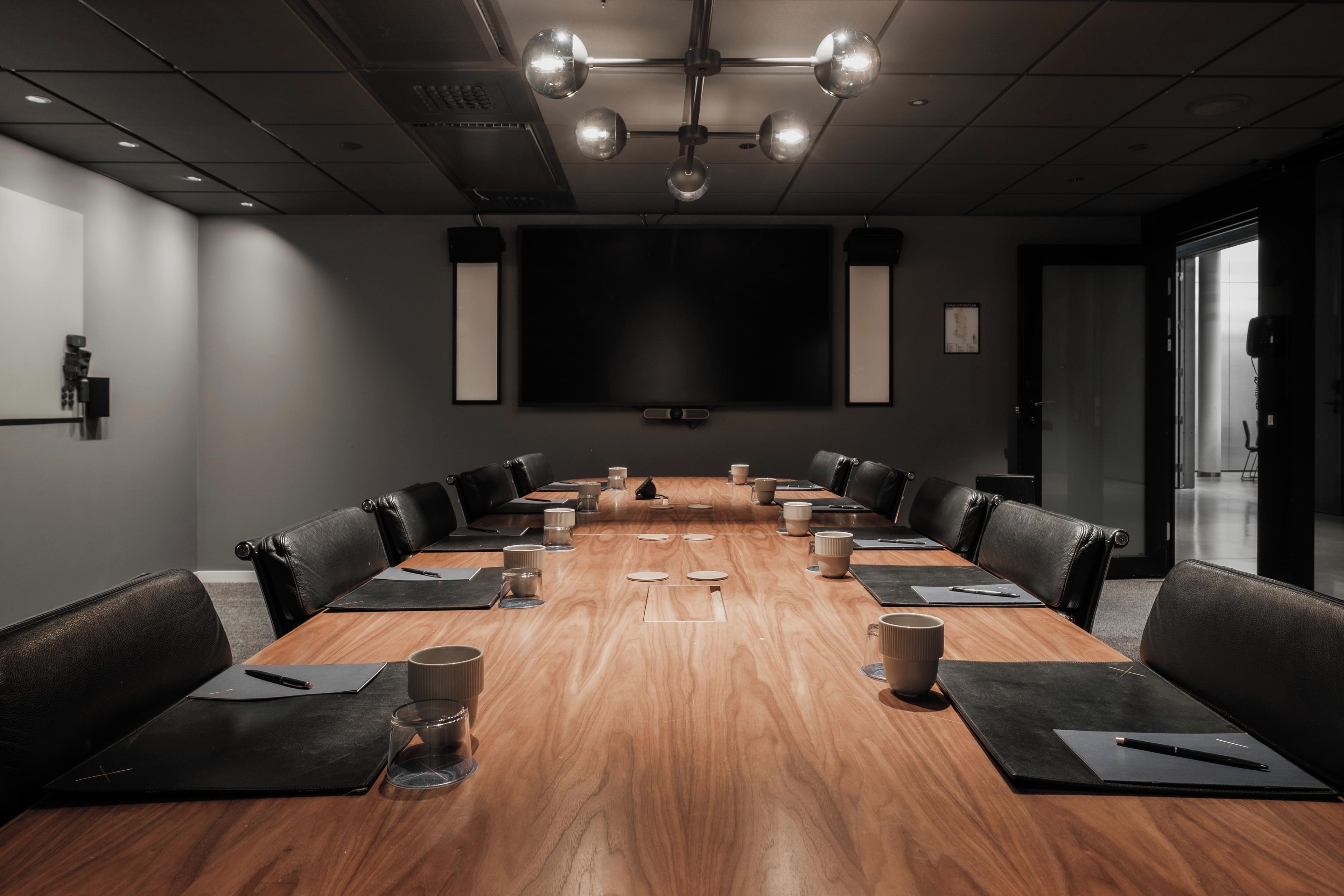 A conference room with a long table, black leather chairs, and a large screen at the front.