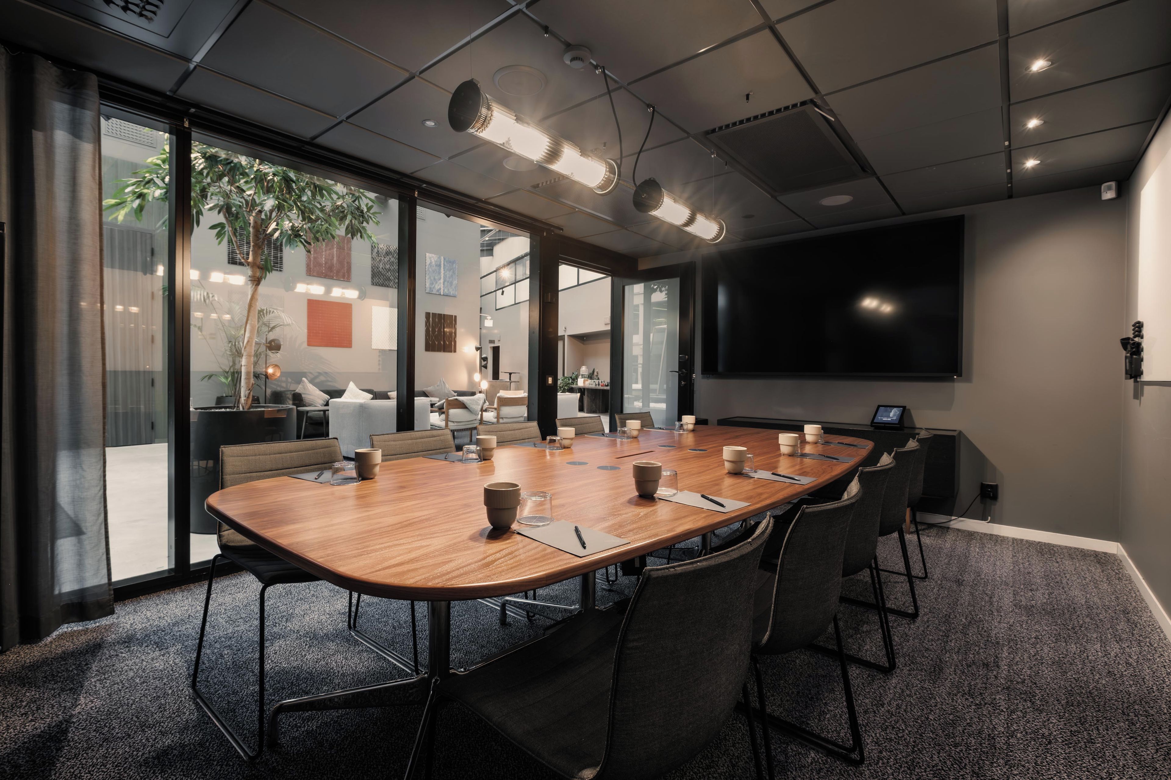 Ett konferensrum med ett långt bord, stolar, en projektorduk och ett stort fönster med utsikt över byggnader och grönområden.