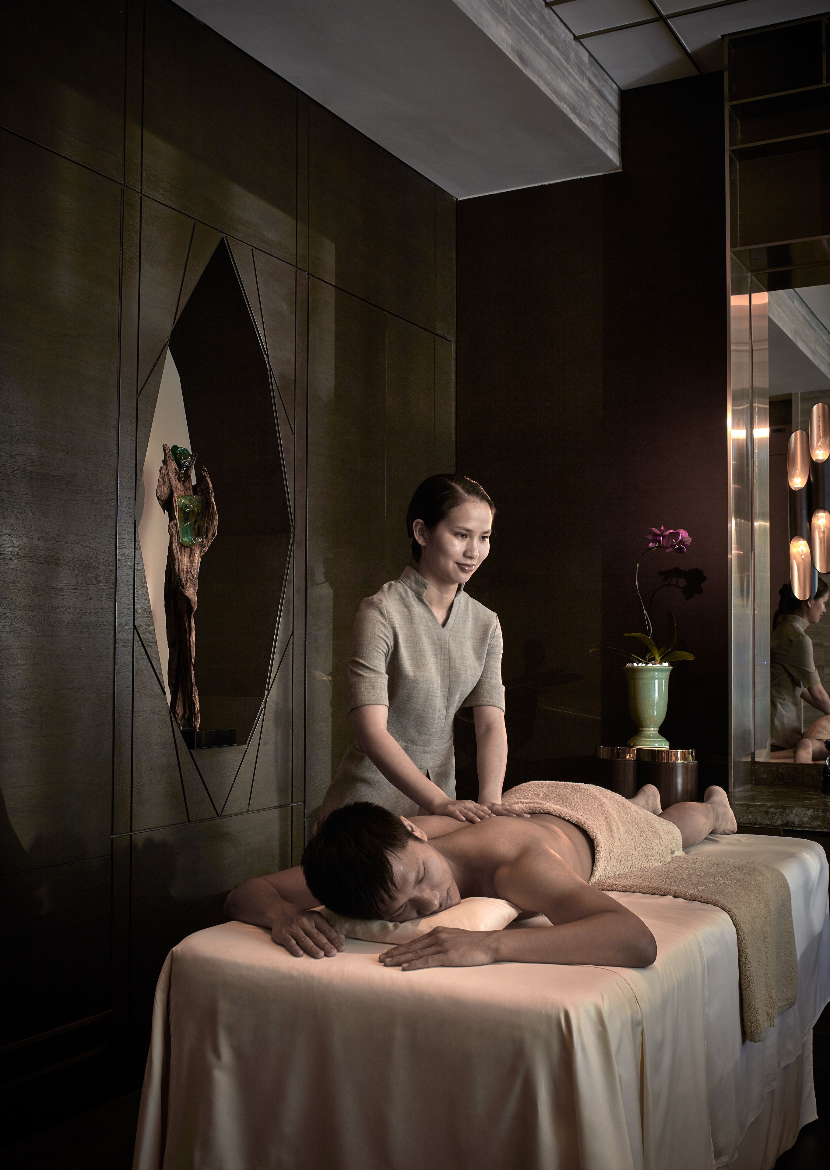 A massage therapy session in a spa setting with a wooden wall, a potted plant, and a mirror reflecting the room.