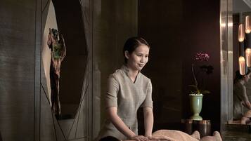 A massage therapy session in a spa setting with a wooden wall, a potted plant, and a mirror reflecting the room.