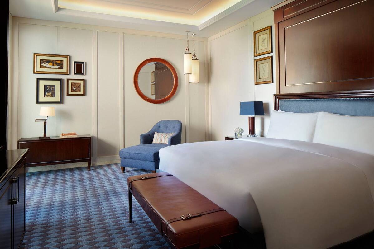 A hotel room with a large bed, a round mirror, a blue armchair, a wooden bench, a desk with a lamp, and framed pictures on the wall.