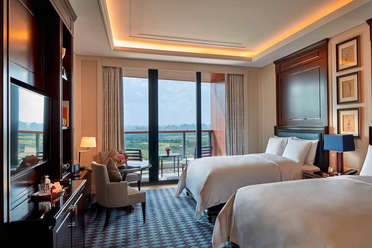 A hotel room with two beds, a desk with a chair, a TV, and a balcony with a view.