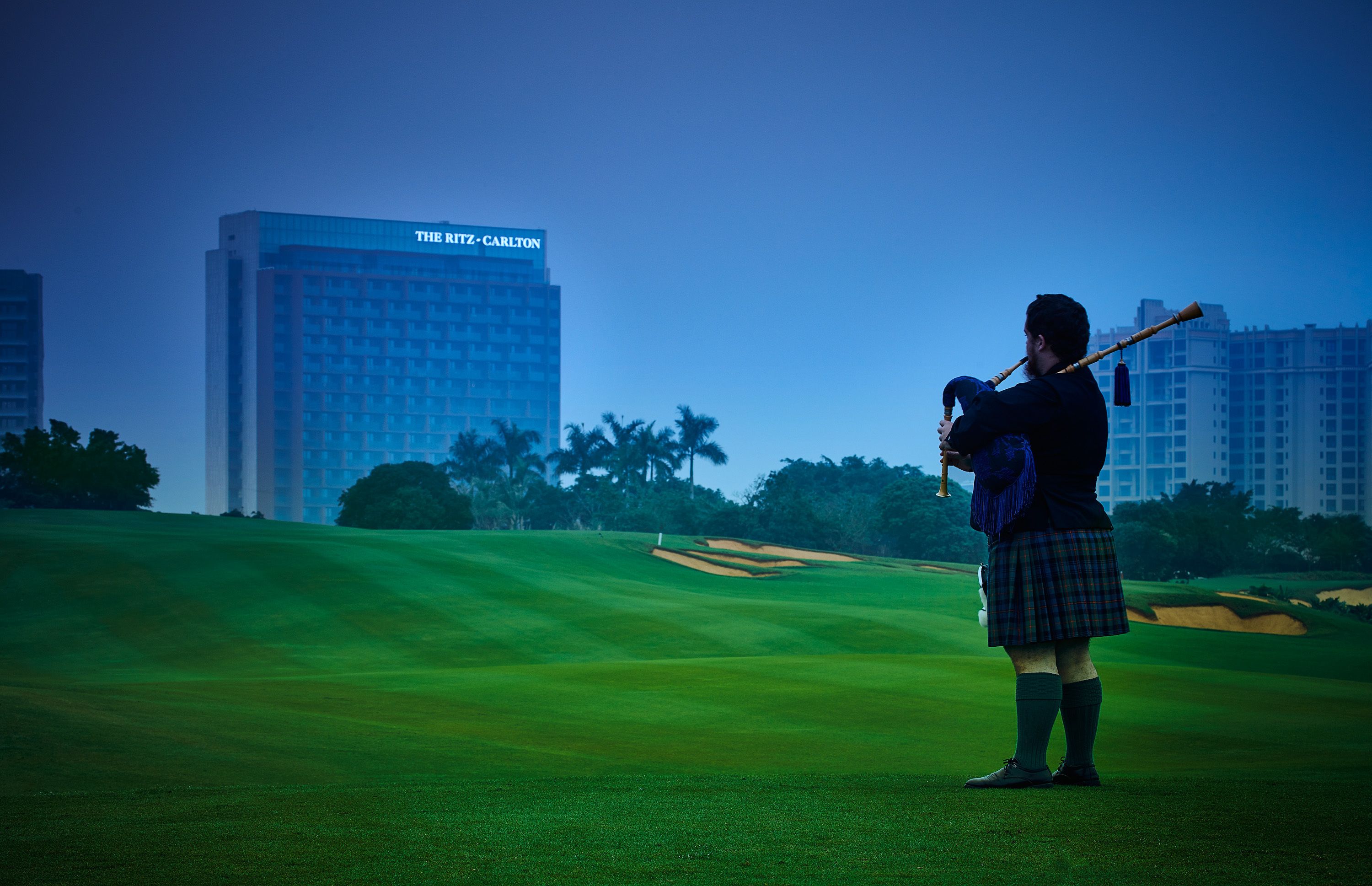 Une personne vêtue d’un costume traditionnel joue de la flûte sur un terrain de golf, avec un bâtiment moderne en arrière-plan.