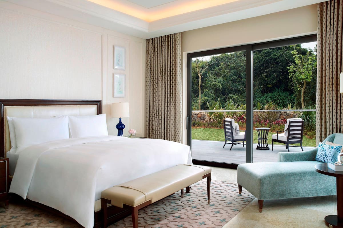 A hotel room with a large bed, a bench, a chair, a small table, and a view of the garden through a sliding glass door.