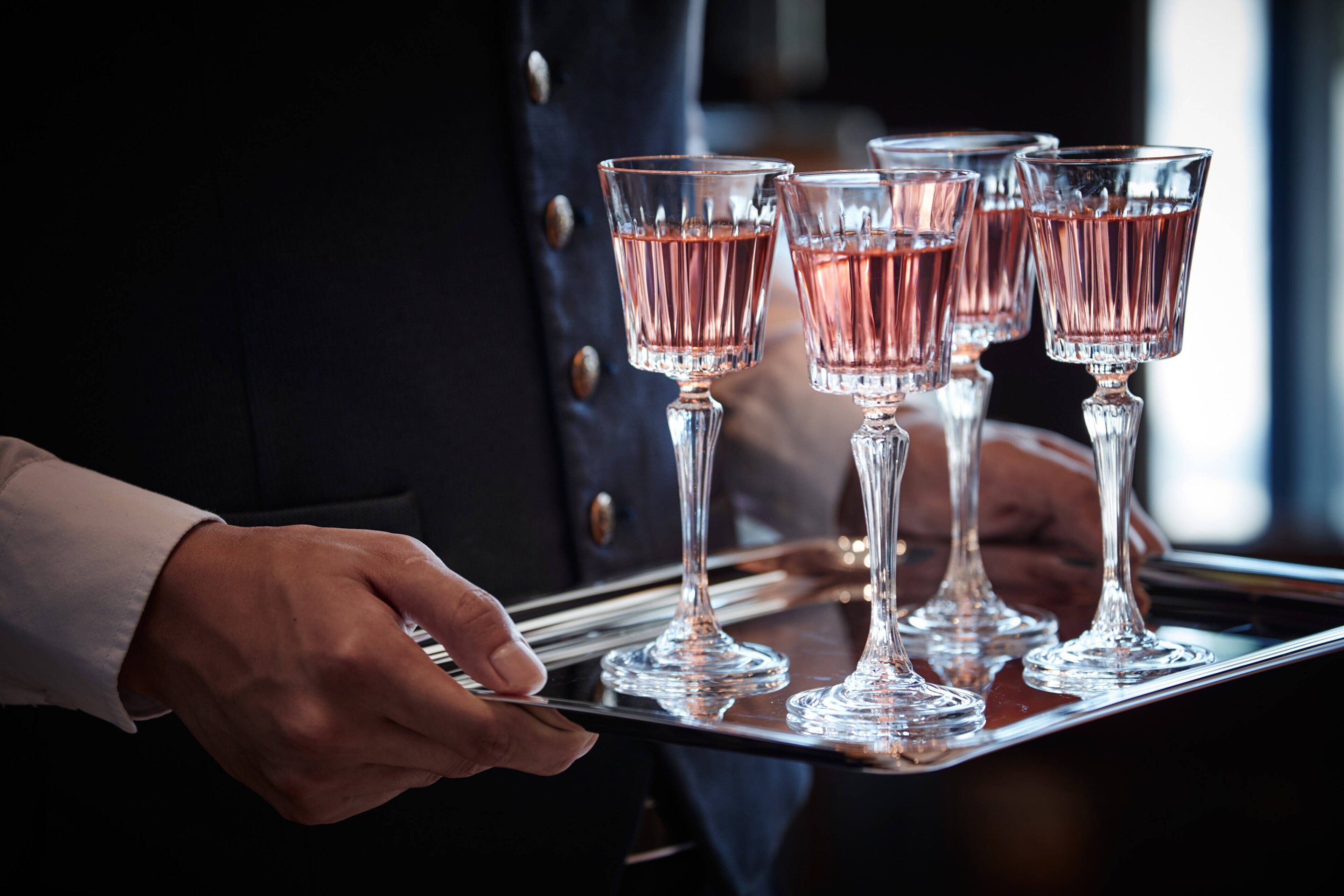 A tray with four glasses of pink liquid, held by a person in a dark uniform.