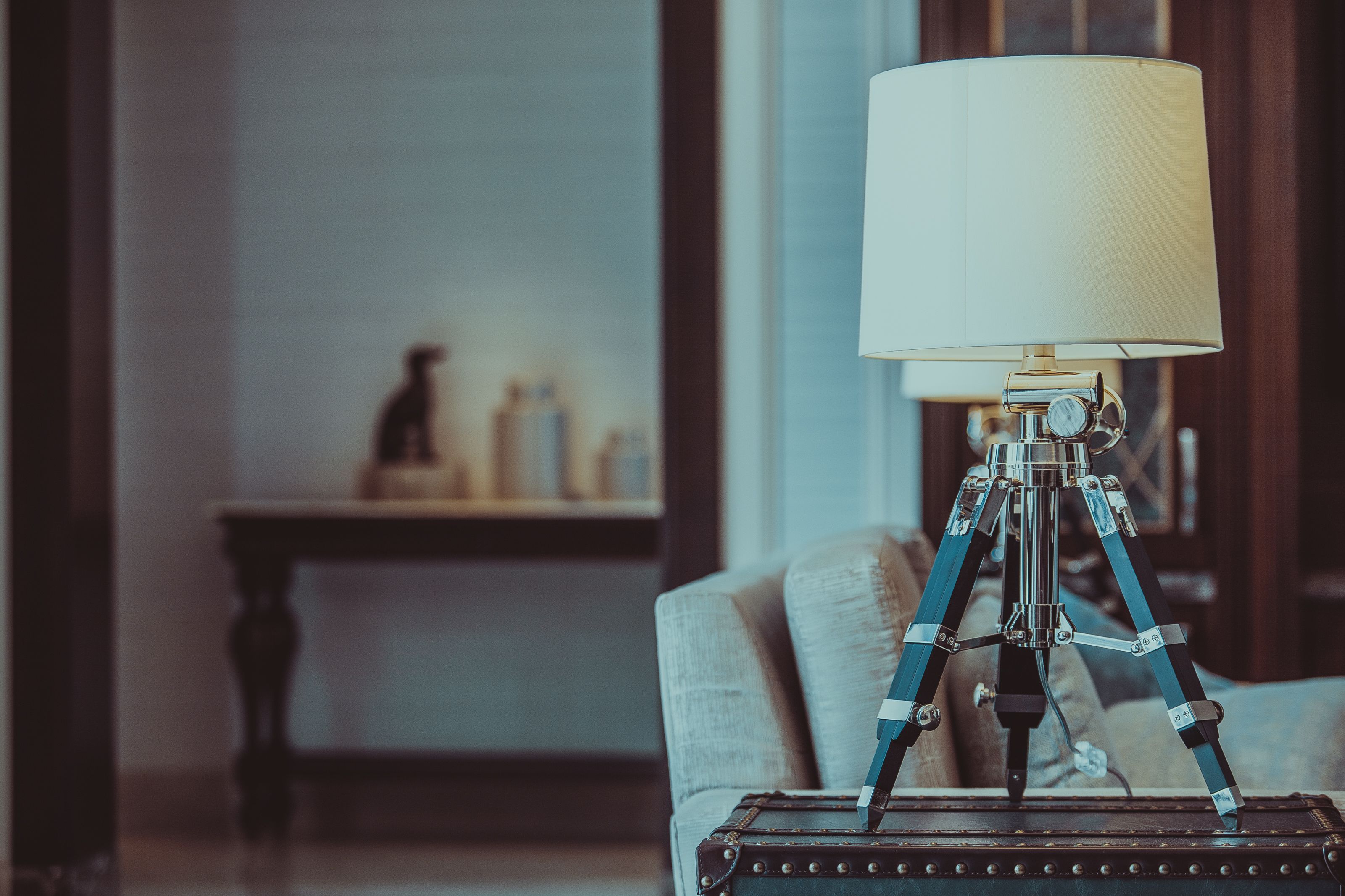 A vintage-style lamp on a tripod stand, a suitcase, and a sofa in a room with a wooden table and a shelf in the background.