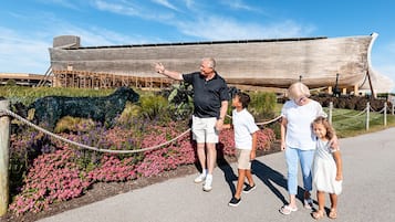 Uma família caminhando perto de um modelo grande de navio, com uma barreira de corda e canteiros de flores.