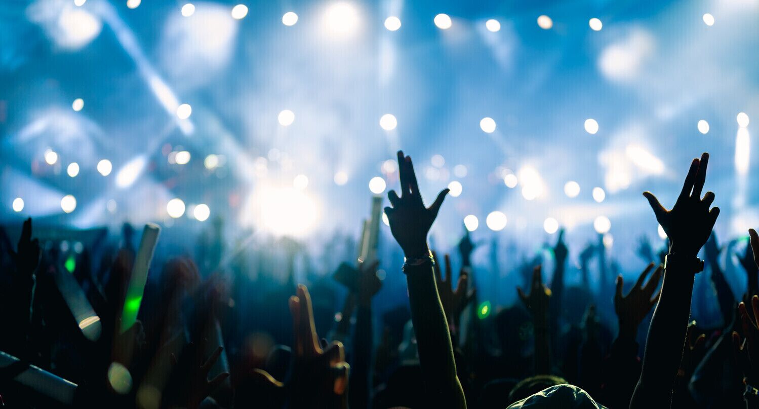 A concert with a crowd of people raising their hands, illuminated by stage lights.