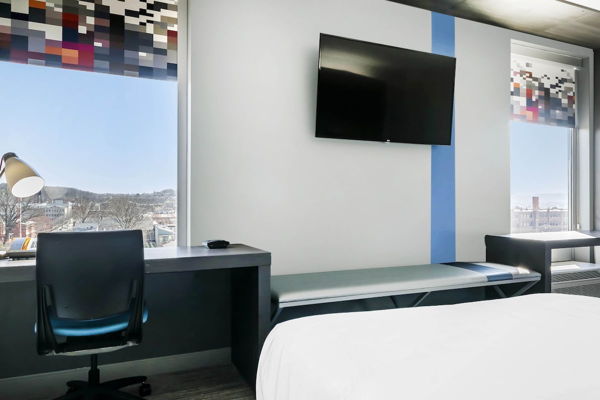 A hotel room with a bed, a desk, a chair, a TV, and a window with a view of buildings.