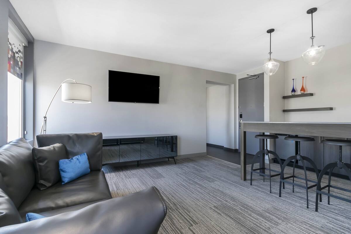 Una sala de estar moderna con un sofá de cuero, un televisor de pantalla plana, una zona de comedor con taburetes de bar y un ventanal grande.