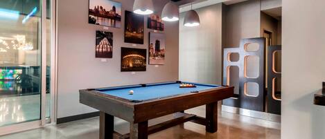 Una mesa de billar con superficie de fieltro azul, rodeada por un interior moderno con iluminación colgante y fotografías de la ciudad en la pared.