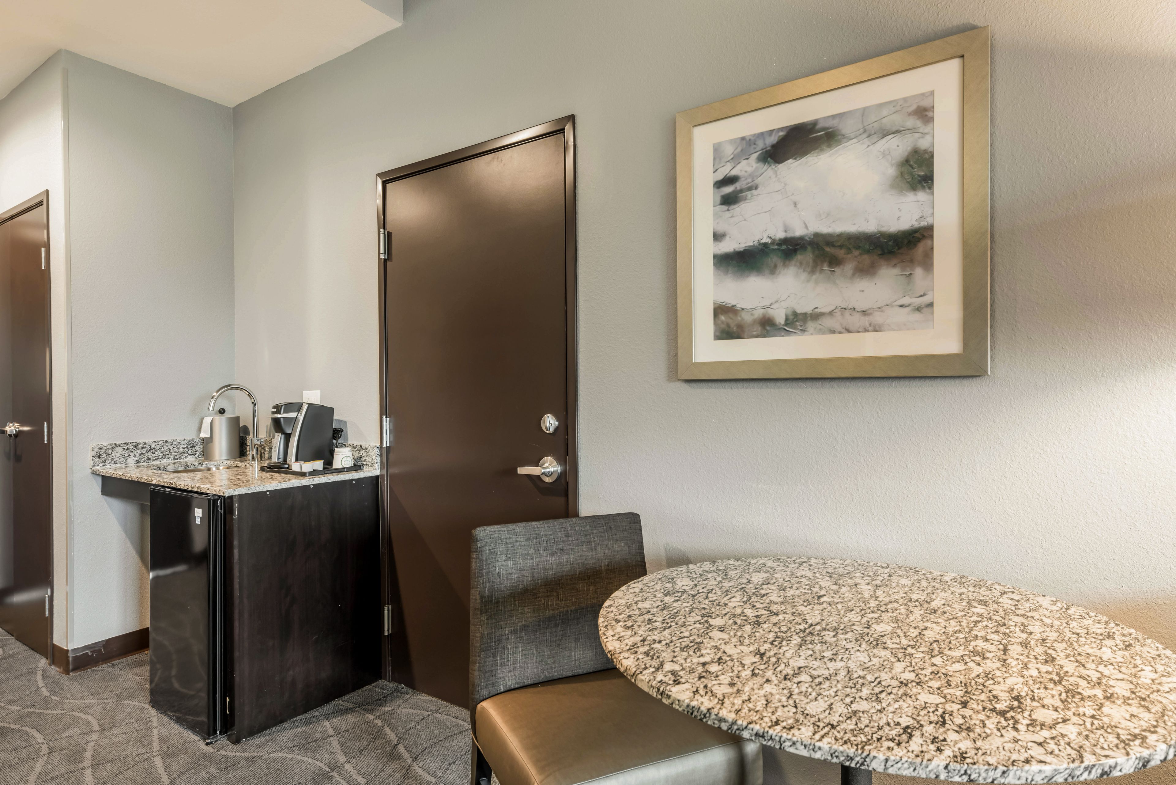 Une chambre d’hôtel dotée d’une kitchenette, d’une table ronde avec un plateau en marbre, d’un fauteuil recouvert d’un tissu gris et d’un tableau abstrait encadré accroché au mur.