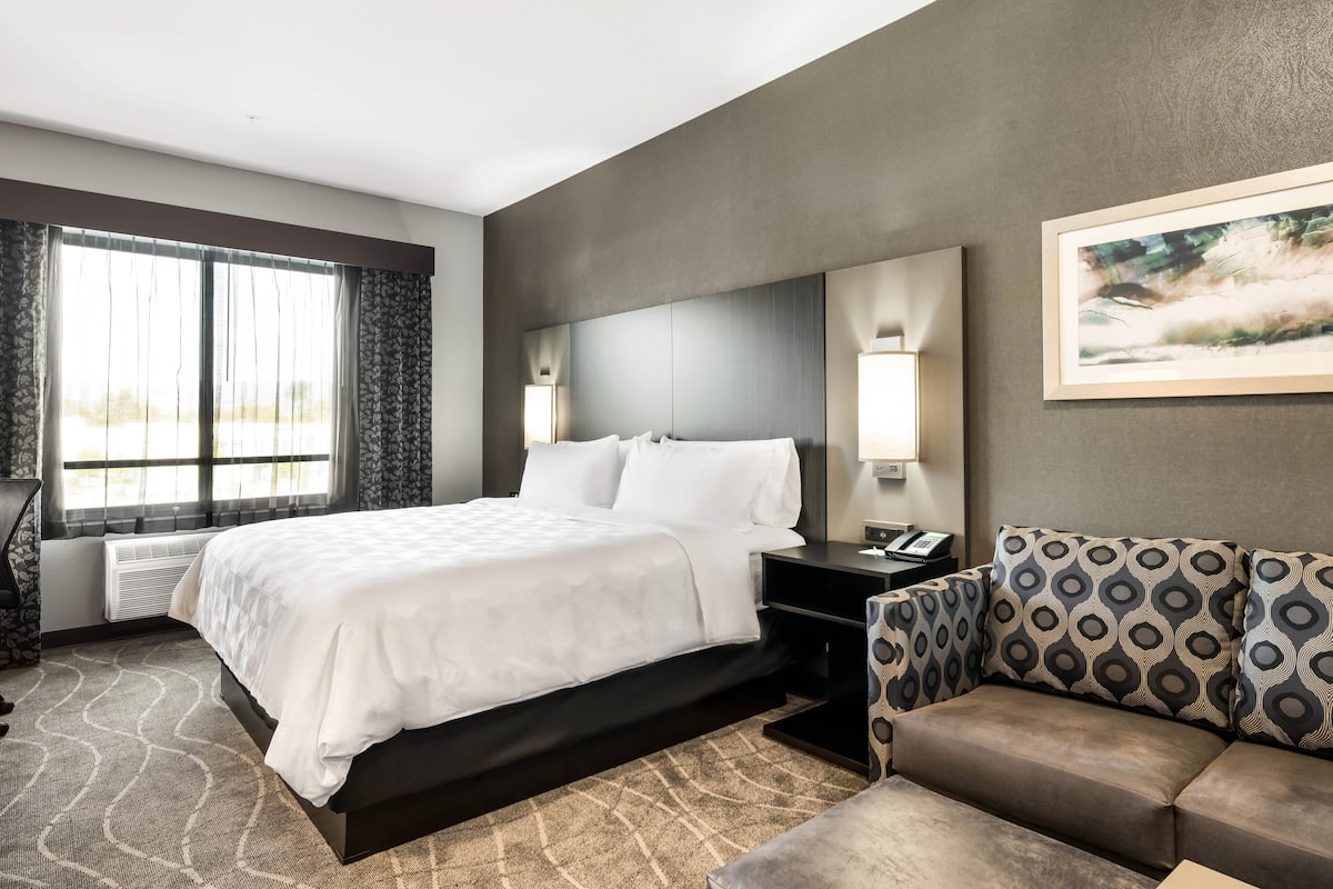 A modern hotel room with a large bed, a sofa, a desk, and a window with curtains.