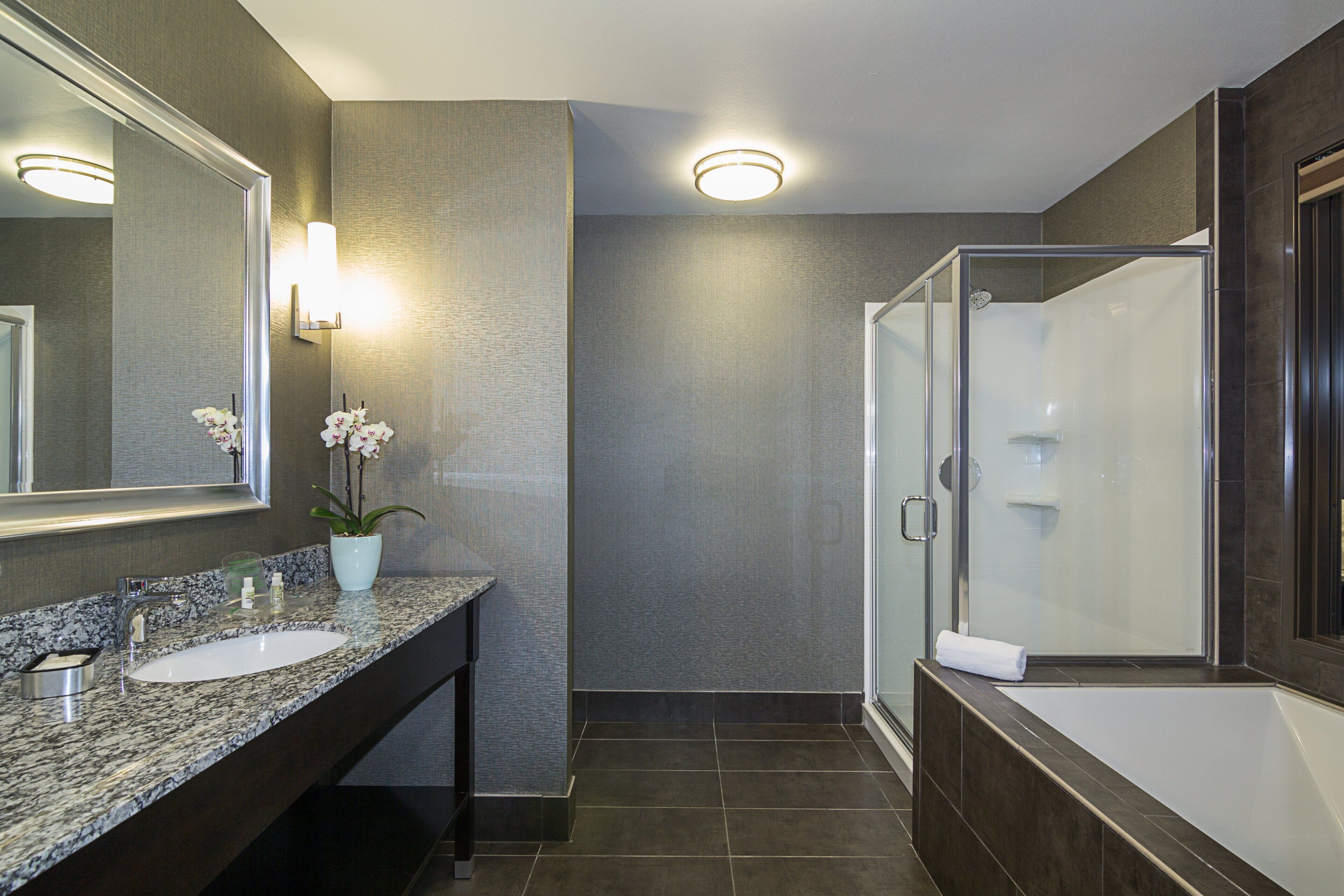 A modern bathroom with a granite countertop, a large mirror, a potted orchid, a glass-enclosed shower, and a dark tiled floor.