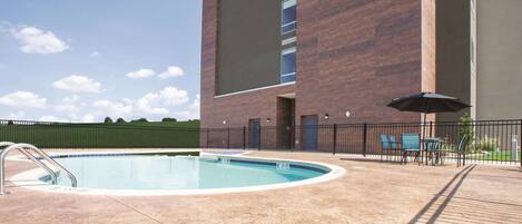 A swimming pool with a metal ladder, surrounded by a brick building with large windows and a covered patio area with outdoor furniture.
