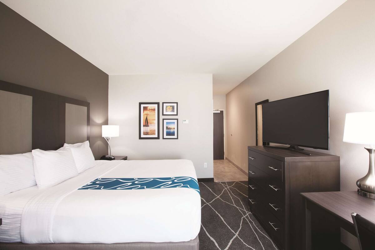 A hotel room with a bed, a desk, a TV, a dresser, and framed pictures on the wall.