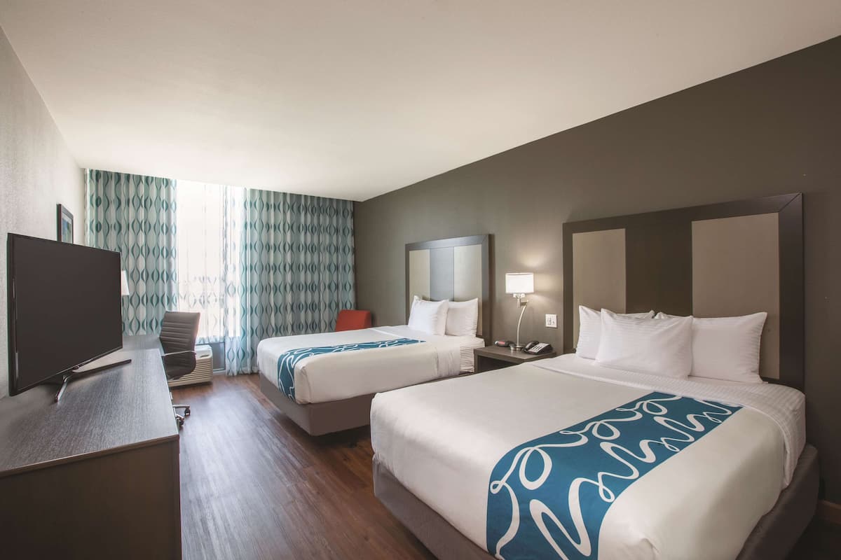A hotel room with two beds, a desk with a TV, a lamp, and a window with patterned curtains.