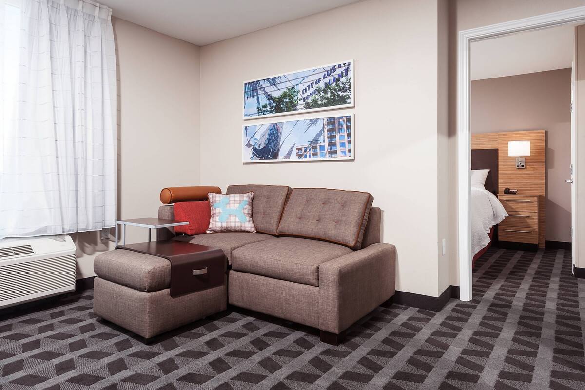 A hotel room with a sectional sofa, a small table, a wall-mounted lamp, and two framed pictures.