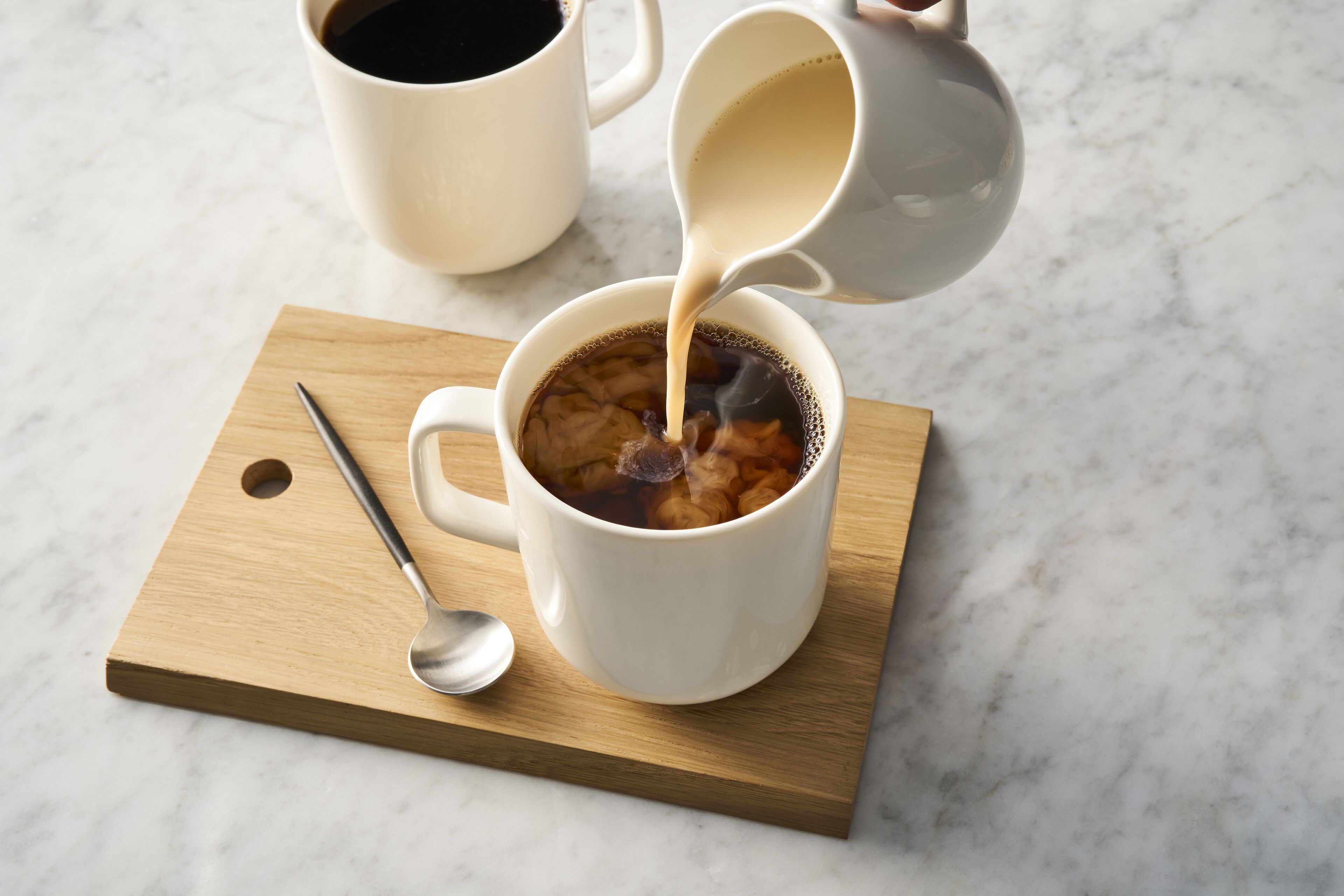 A white mug on a wooden board with a spoon, and a white mug being filled with a dark liquid.