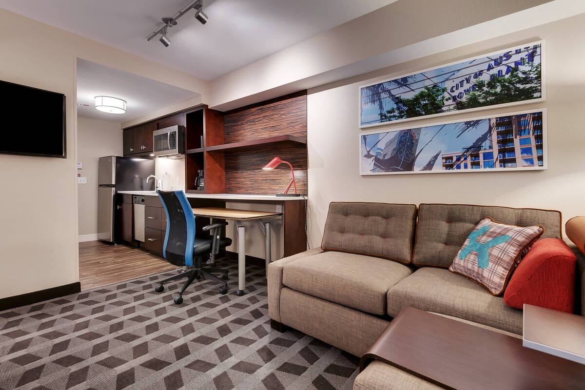 A compact hotel room with a kitchenette, a sofa, a desk, and a TV.