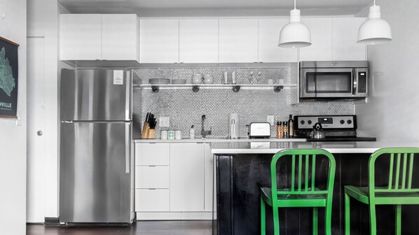 A modern kitchen with a stainless steel refrigerator, a microwave, a kettle, and a toaster. There are two green chairs and a white countertop. - The Sobro Guest House by AvantStay (Nashville)
