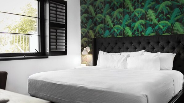 Una cama bien tendida con sábanas blancas, un cabecero oscuro tapizado y una ventana con contraventanas negras, además de una pared decorada con un diseño de hojas tropicales.