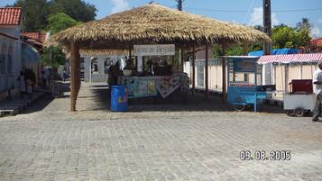 Un étal de marché rustique, avec un toit de chaume, une enseigne indiquant « ENTRO DO ARTESÃO » et une poubelle bleue.