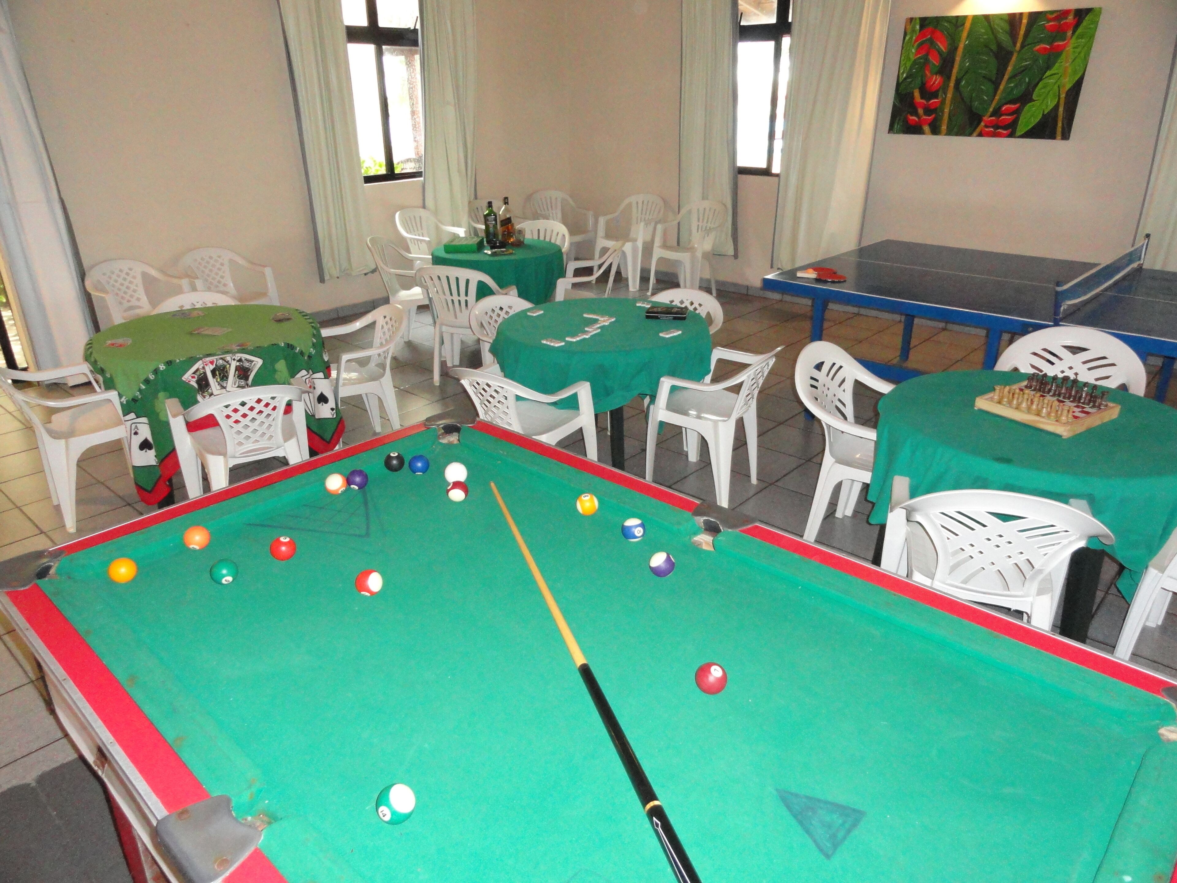 A pool table with balls and a cue stick, surrounded by white plastic chairs and tables with green tablecloths.