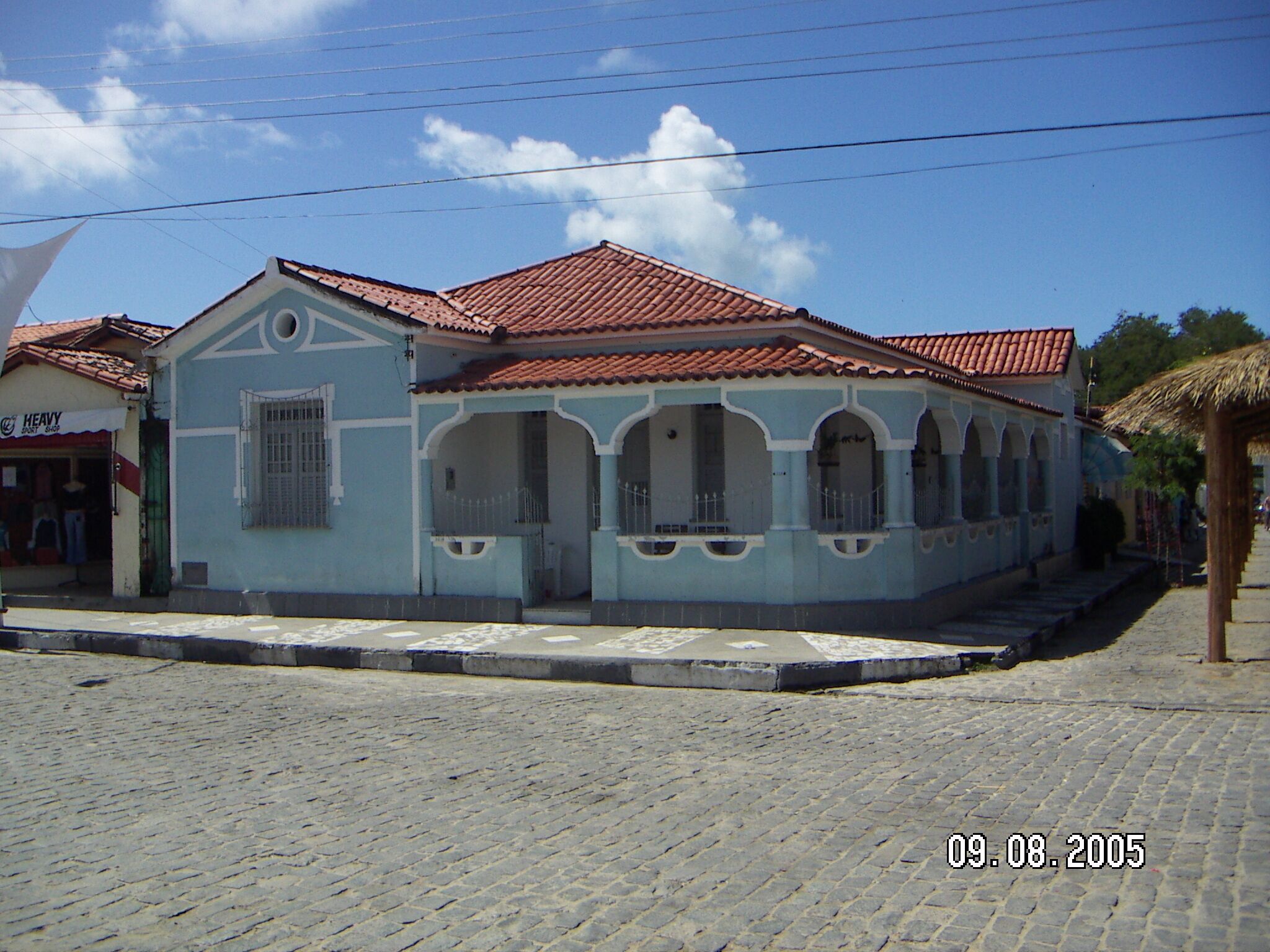 A light blue building with a red-tiled roof, a small porch, and a shop on the ground floor.
