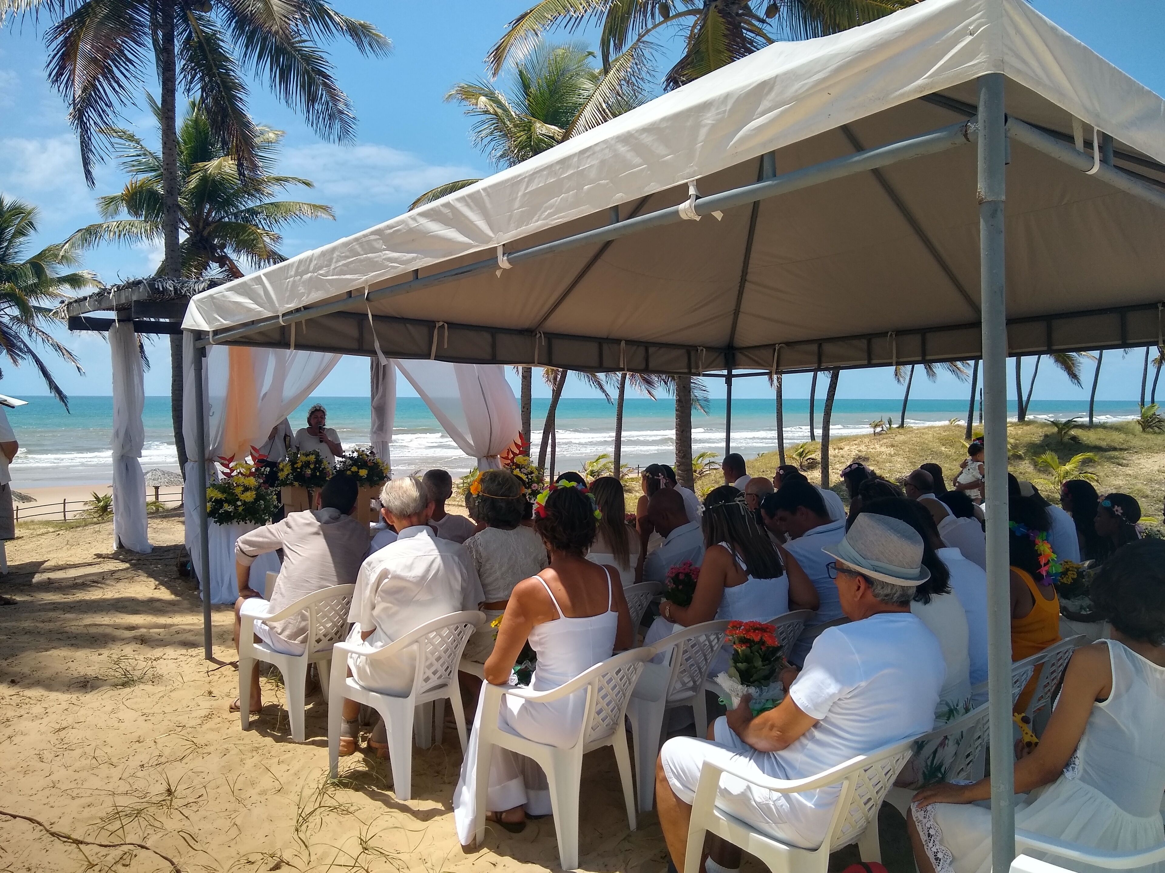 A beach wedding ceremony under a tent with guests seated on white plastic chairs, palm trees, and the ocean in the background.