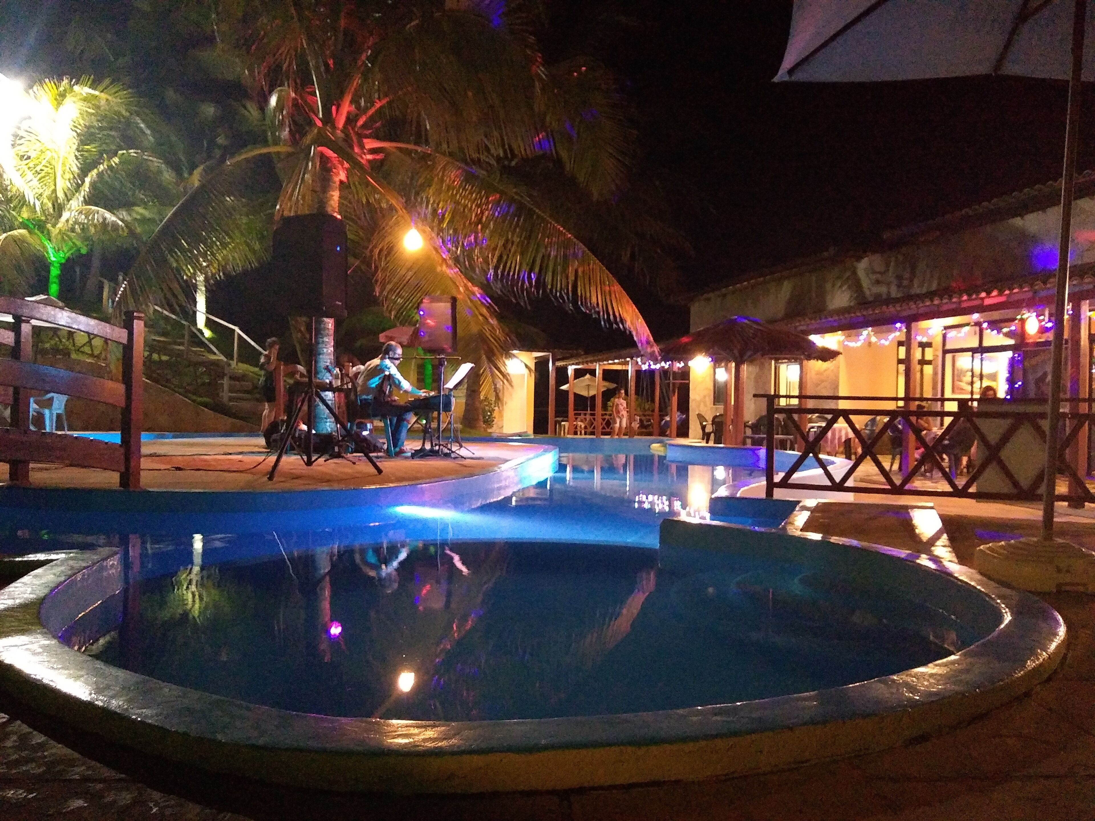 A nighttime pool area with a stage for live music, palm trees, and a building with outdoor seating.