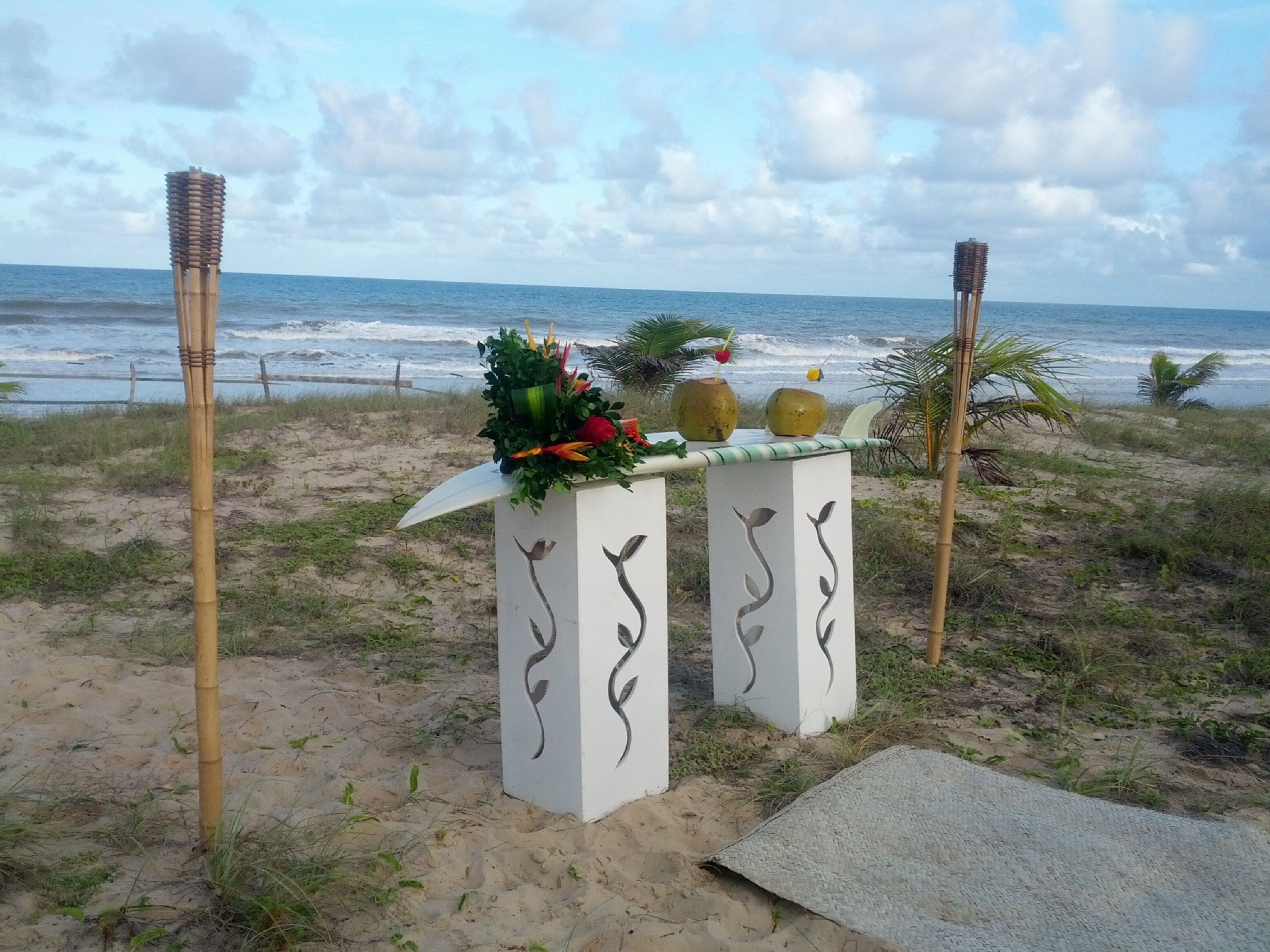Um altar à beira-mar com arranjo floral e dois vasos em formato de coco, com o oceano e um céu nublado ao fundo.