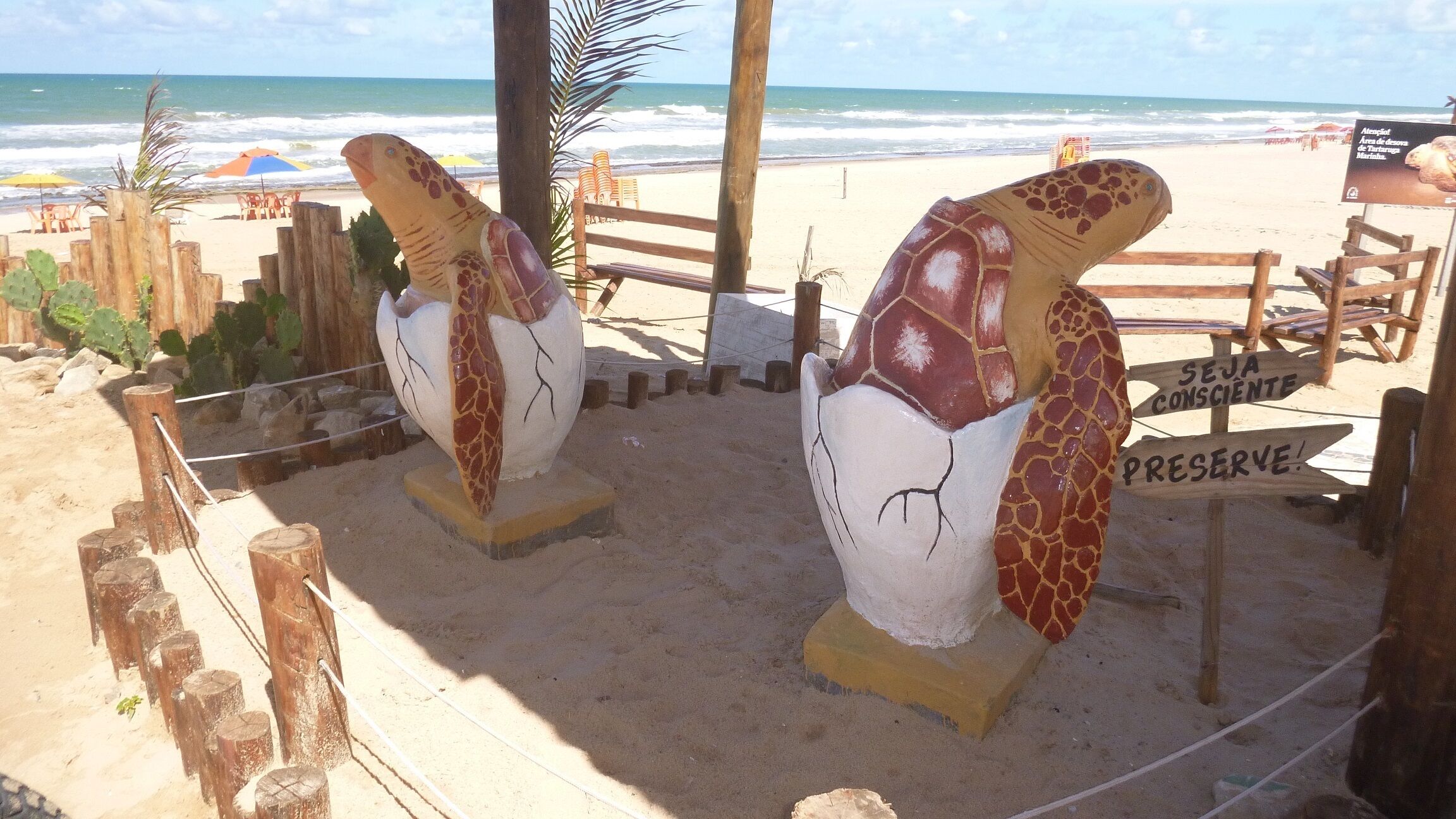 A beach with two large sculptures of a turtle and a tortoise, a wooden fence, and a sign that says "SEJA CONSCIENTE PRESERVE!".