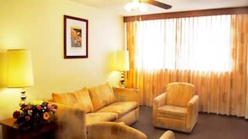 Habitación de hotel con sofá, sillón, dos lámparas, ventilador de techo y ventana con cortinas.