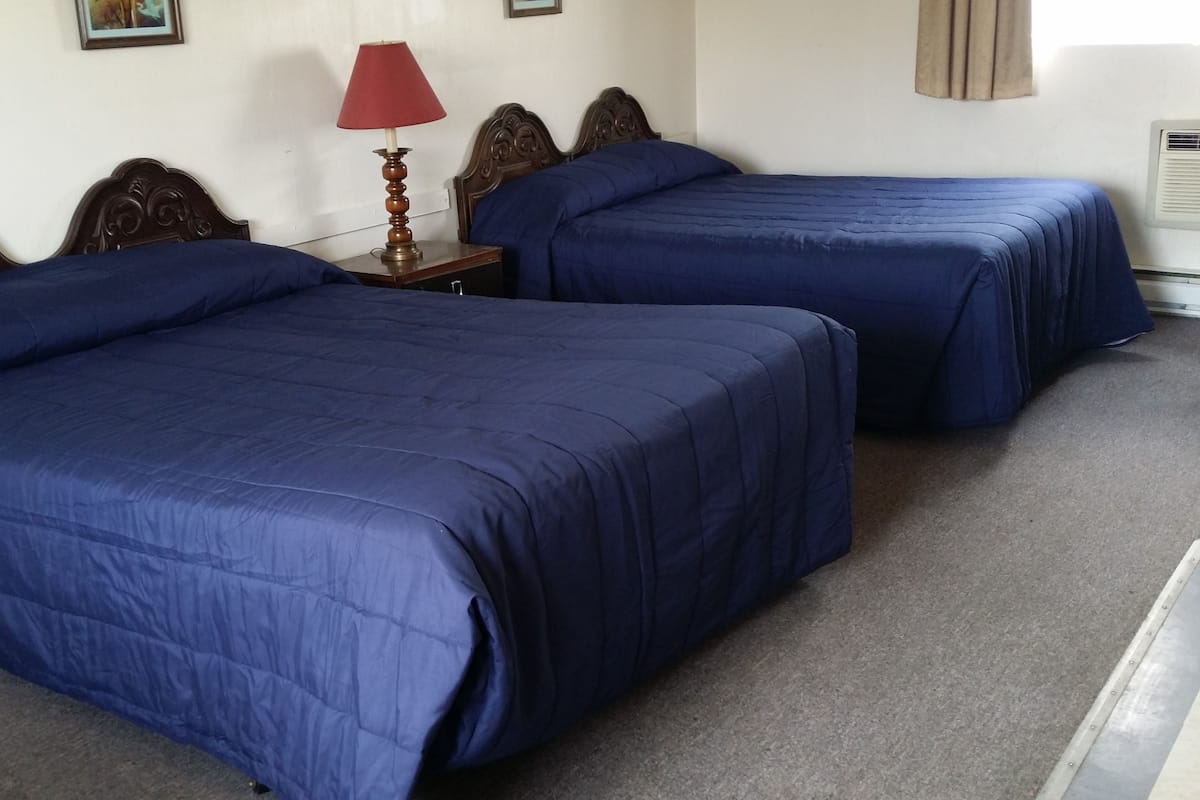 Ein Hotelzimmer mit zwei Betten, jeweils mit blauer Bettdecke, einem hölzernen Kopfteil, einem Nachttisch mit roter Lampe und einer wandmontierten Klimaanlage.