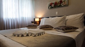 Cama bem arrumada com lençóis brancos e almofadas decorativas, um quadro de flores na parede e uma luminária em uma mesa de cabeceira.