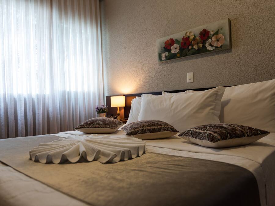 Cama bem arrumada com lençóis brancos e almofadas decorativas, um quadro de flores na parede e uma luminária em uma mesa de cabeceira.
