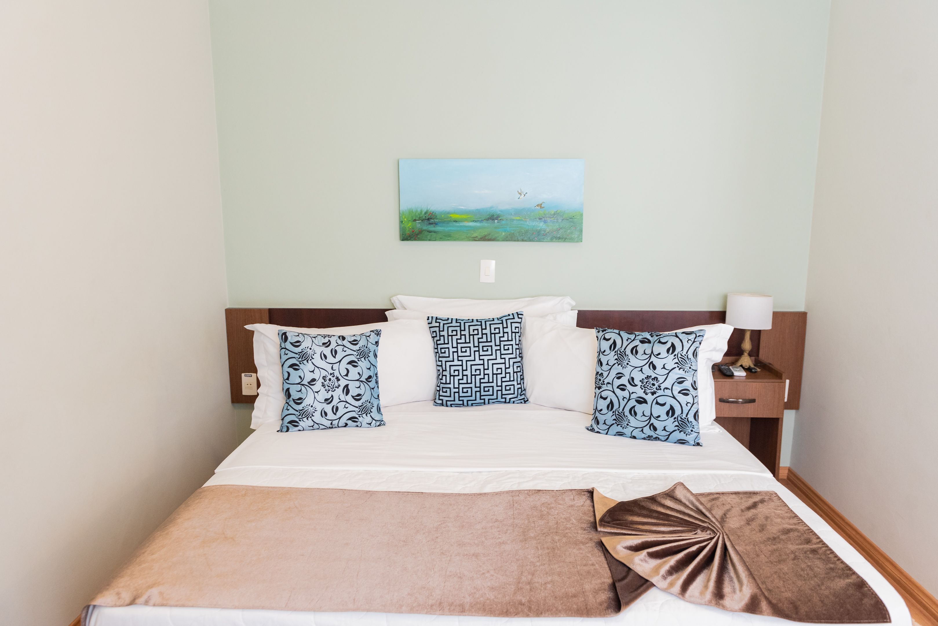 A neatly made bed with patterned pillows, a brown throw, and a painting of a landscape on the wall.