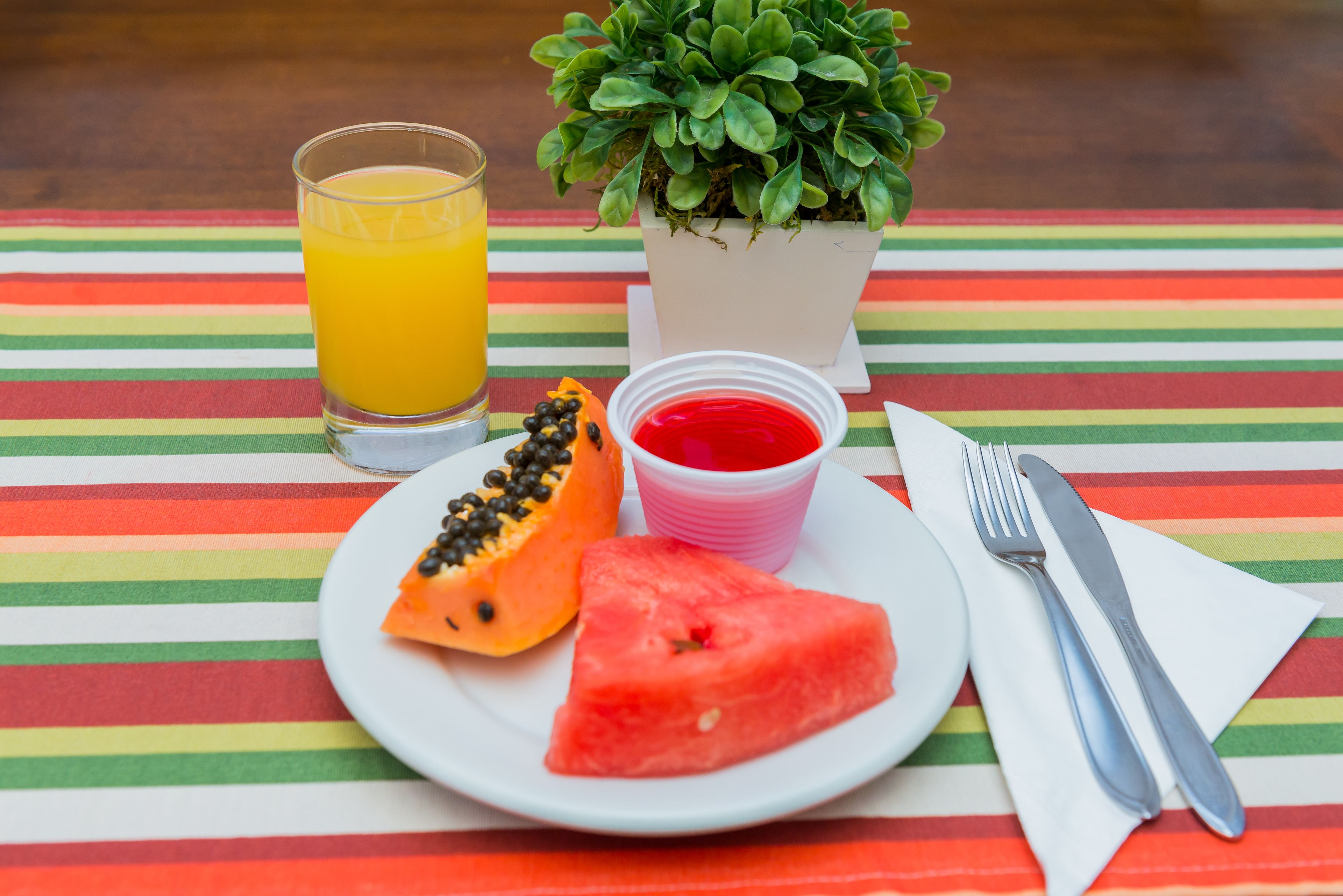 A plate with a slice of watermelon and a half-cut papaya, a glass of orange juice, and a small potted plant on a striped tablecloth.
