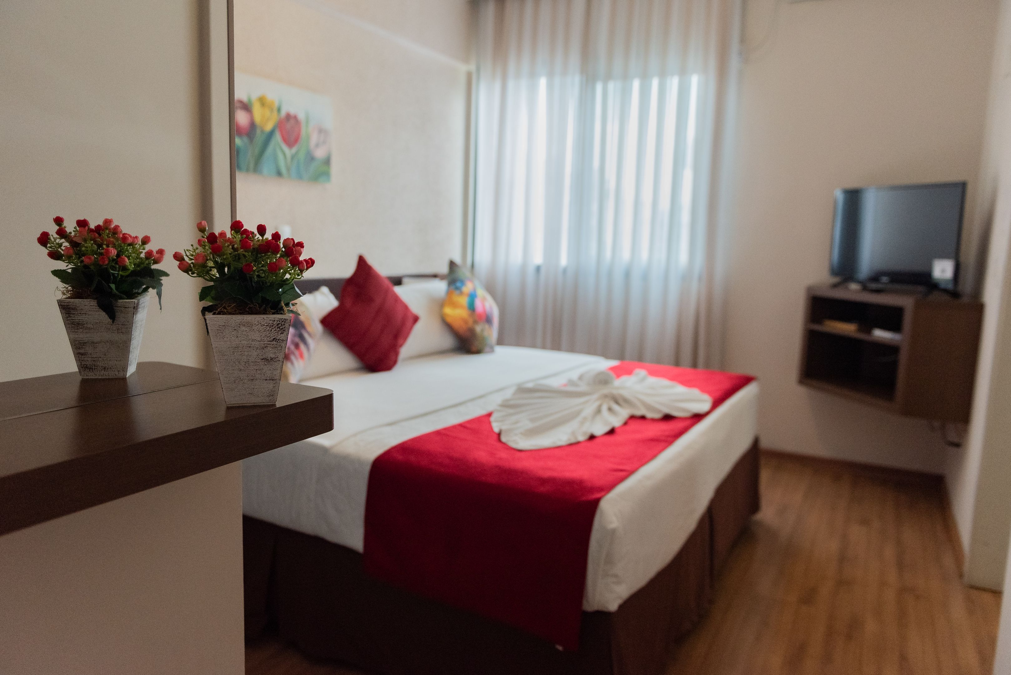 A hotel room with a bed, a red blanket, two potted plants, a red pillow, a television on a stand, and a painting on the wall.