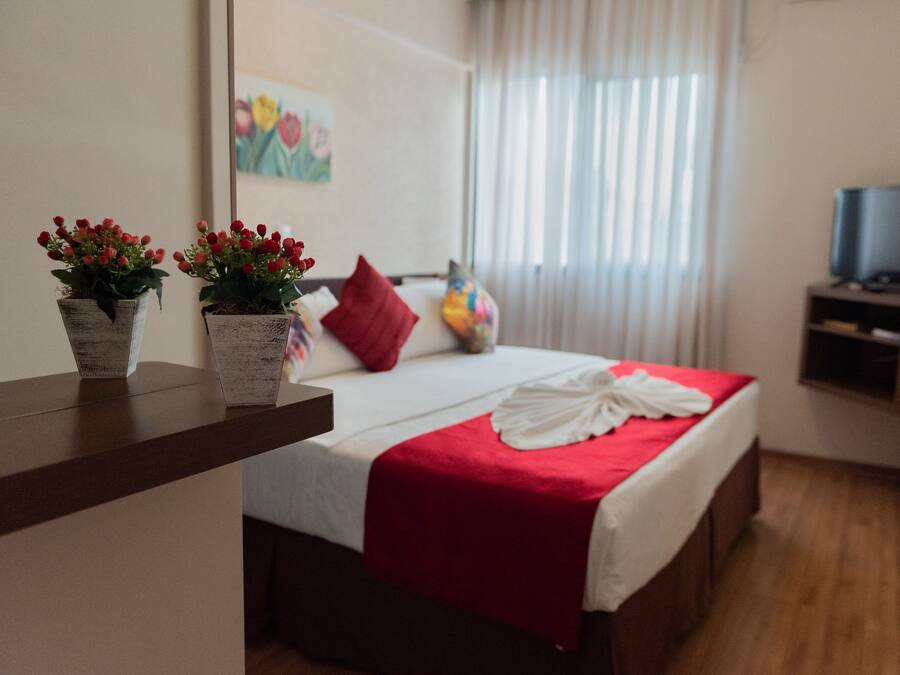 Una habitació d'hotel amb un llit, una manta vermella, dues plantes en testos, un coixí vermell, una televisió sobre un suport i un quadre a la paret.