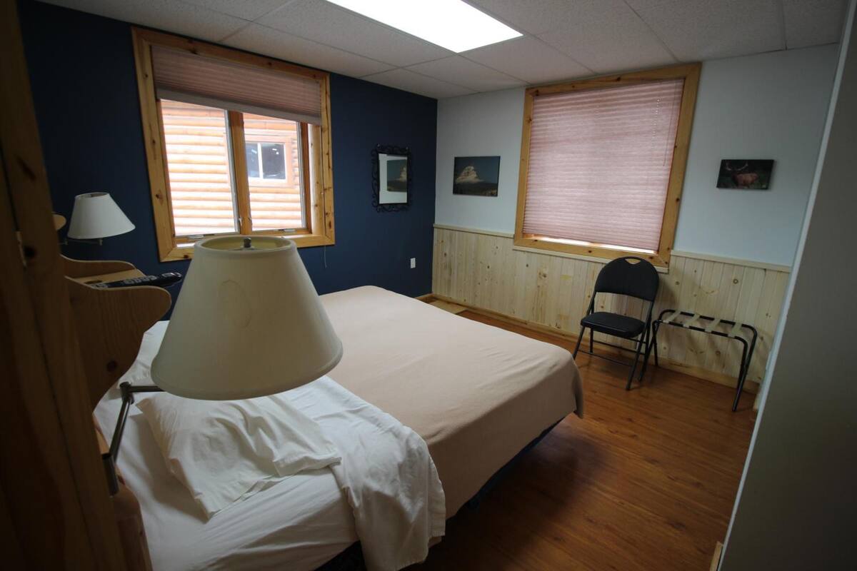 Une chambre à coucher avec un lit, une chaise, une lampe et deux fenêtres équipées de stores.