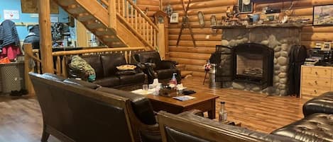 A log cabin interior with a stone fireplace, leather furniture, a wooden staircase, and mounted animal heads.