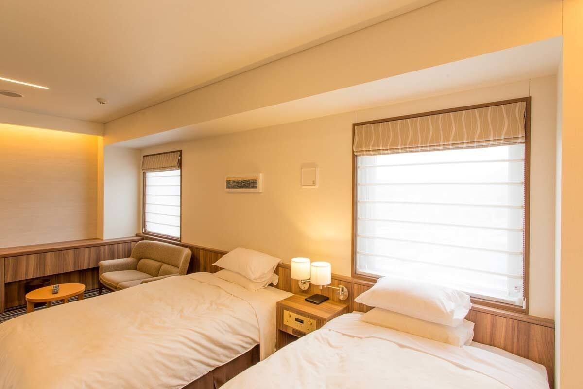 Hotelkamer met twee bedden, een klein tafeltje, een stoel en een raam met zonwering.