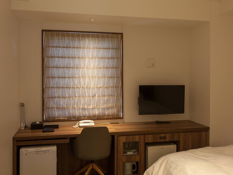 Una habitación de hotel con un escritorio de madera, televisor, cama y ventana con persianas.