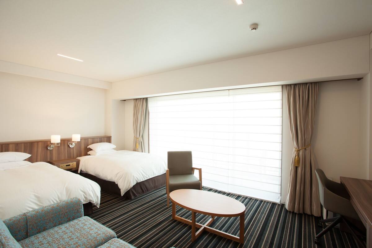 Hotelkamer met twee bedden, een klein rond tafeltje, een bank, een bureau en een groot raam met gordijnen.