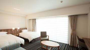 A hotel room with two beds, a small round table, a sofa, a desk, and a large window with curtains.