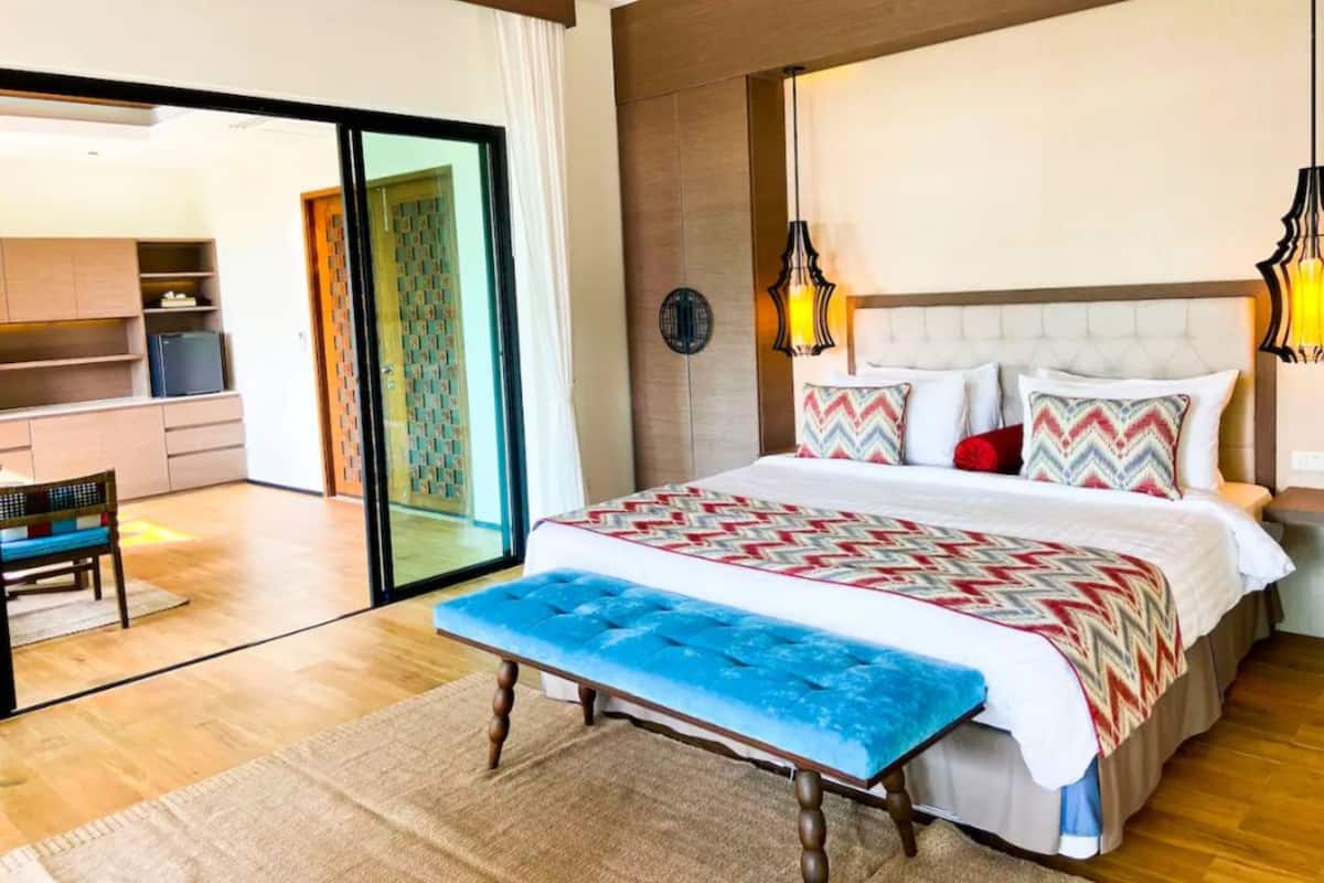 A modern hotel room with a large bed, a bench, a desk, and a sliding glass door leading to a balcony.