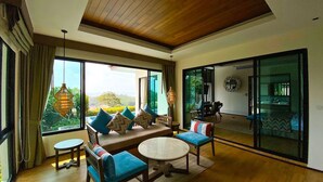 A spacious living area with a wooden ceiling, large windows offering a view of greenery, a sofa with cushions, a round coffee table, and a small side table. - Silla Villa Private Pool & Sea View (Karon)