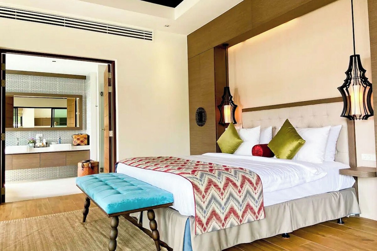 A bedroom with a large bed, a bedside table, a nightstand, a bench, a mirror, and a window.